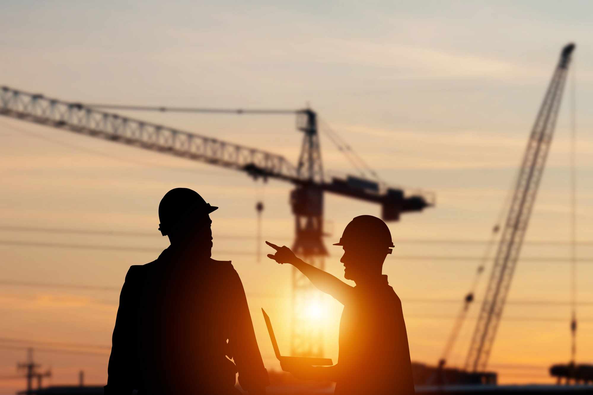 Silhouettes of two construction workers working on a construction site at sunset with the sun and a crane in the background.