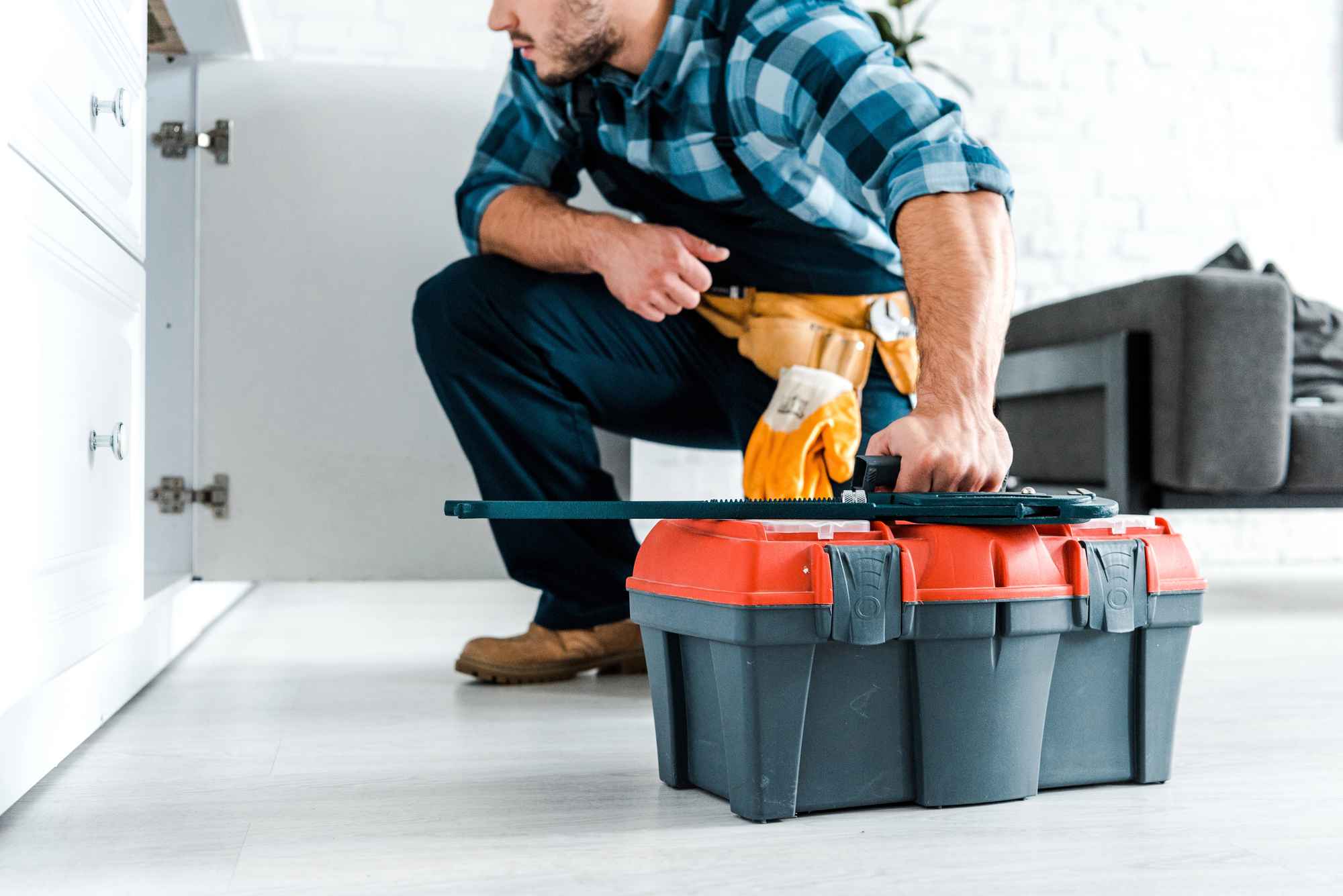 A handyman with a toolbox looking under a sink.