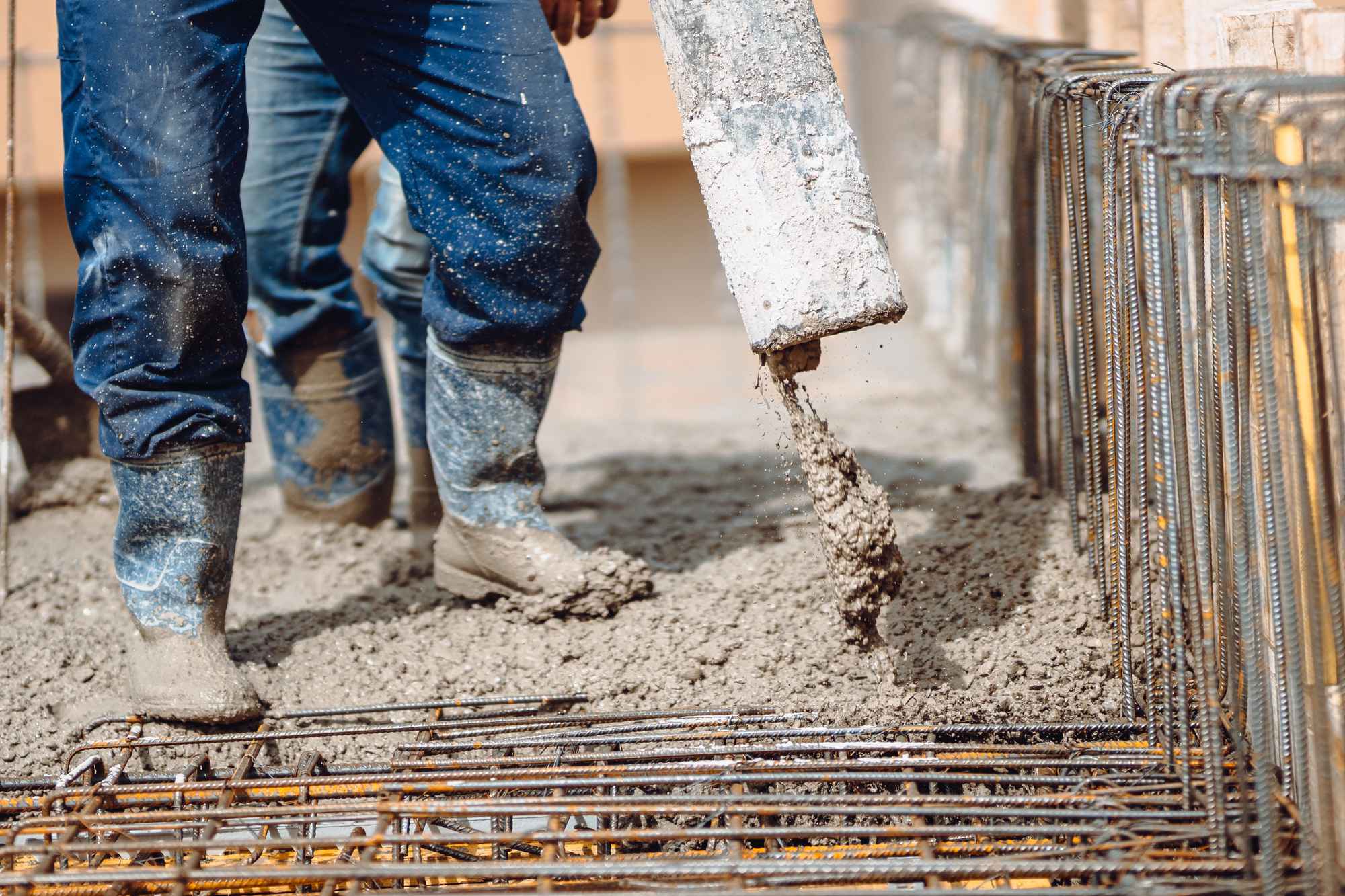 A construction worker in blue jeans and rubber boots pouring concrete into a foundation with rebar.